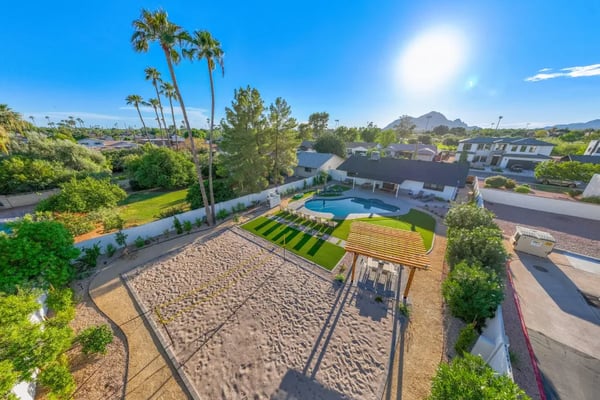 Aerial view of desert home with sand volleyball court, pool, striped lounge area, palm trees, and mountain backdrop